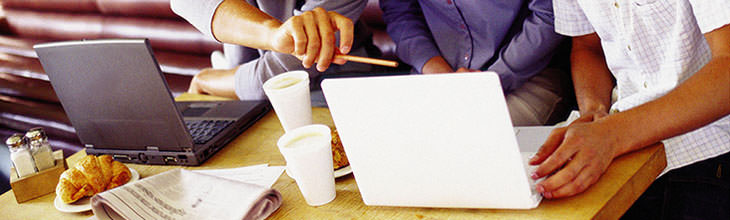 Panoramabild: Moderne Arbeistszenerie: Laptops und Kaffeebecher und Früstücksreste stehen oder liegen herum. Drei Personen beraten sich und tauschen sich am Laptop aus. Die  Körpersprache verrät Angeregtheit und Zielstrebigkeit.